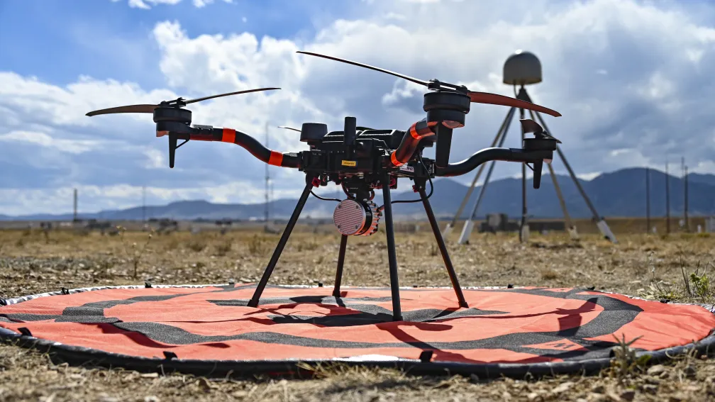 Close photo of a drone on the ground with a GPS station and mountains in the background.