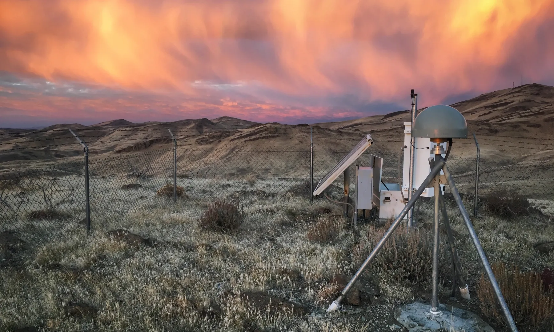 A GNSS station amongst shrubs and a vibrant pink and orange sunset.