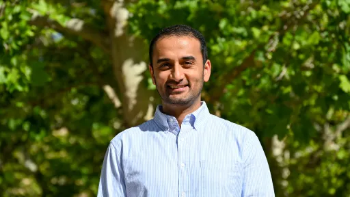 Headshot of Eshanta wearing a button down standing in front of green foliage.