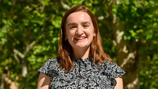 Gabrielle stand in front of green foliage wearing a black and white patterned blouse.