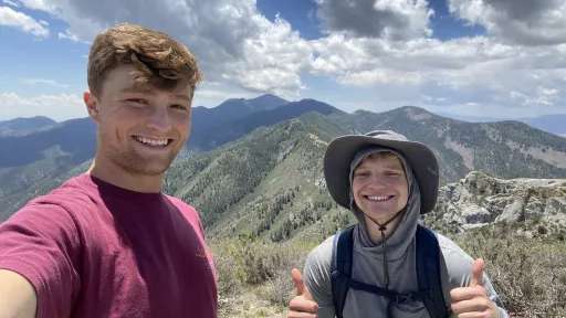 Two smiling people take a selfie photo with mountains in the background, one is giving two thumbs up gestures.