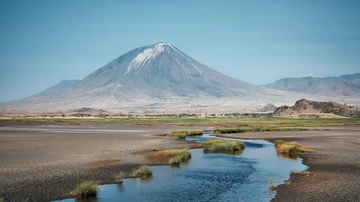 photo of Old Doinyo Lengai, a gray volcano with a patch of streaky white near the summit, foreground is patchy vegetation and a stream