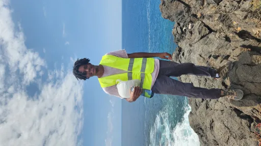Kaetu wearing a high visibility vest and holding a hard hat. Kaetu is standing on rocks that are overlooking the ocean.