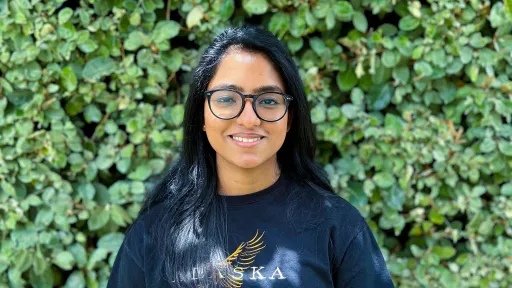 Ann Mariya wearing a black shirt and glasses standing in front of vegetation smiling. 