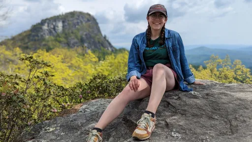 Isabella sitting on a rock in the mountains wearing a blue flannel and a baseball hat.