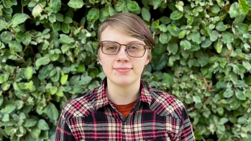 Elliz smiling in front of green foilage wearing a red and black plaid flannel and glasses.