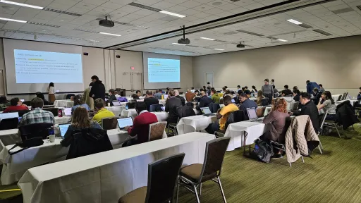 a room full of people at long tables opening up their laptops and looking at projector screens at the front of the room