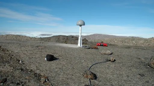 a GPS antenna on a short white metal post affixed to bare bedrock, with white ice marking the horizon in the background