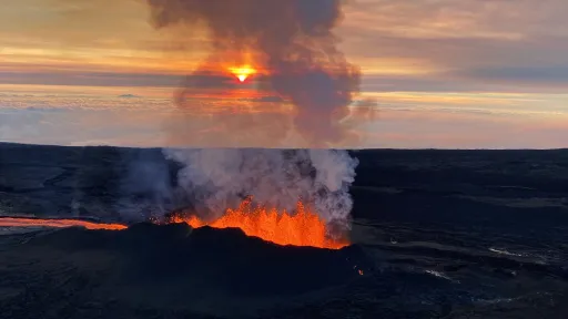 Lava fountaining into the area along a fissure in dark rock. Smoke rises from the lava, reaching up into a sunrise on the horizon.