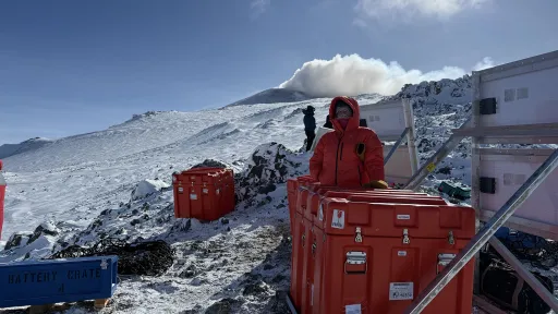 A person stands beside three red cases and two sets of solar panels, with a snowy slope behind and visible steam rising from the peak.