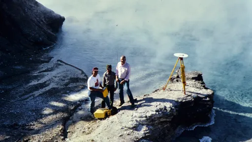 Three geoscientists conduct a GPS survey beside a steaming hydrothermal pool at Yellowstone National Park in 1989. A tripod-mounted GPS antenna is set up on a rocky outcrop. From left to right: Chuck Meertens, Rick Hutchinson, and Bob Smith stand near yellow equipment cases at the water&rsquo;s edge.