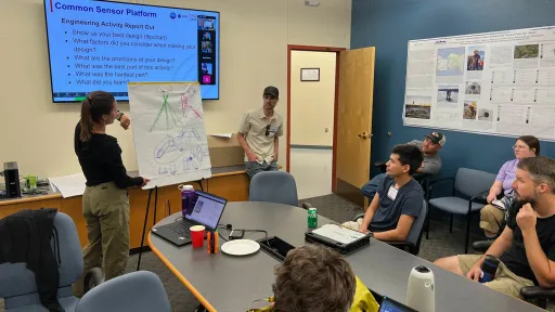 A woman stands at the front of a conference room, presenting a hand-drawn engineering diagram on a flip chart to a seated group. A man stands beside her, and a large screen behind them displays a slide titled &ldquo;Common Sensor Platform: Engineering Activity Report Out,&rdquo; with a list of discussion questions. Several participants watch attentively, with laptops, drinks, and notes on the table.