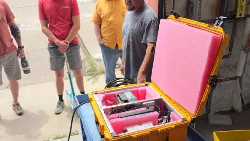 Four engineers surround opened yellow prototype system box that sits on a blue hand cart.