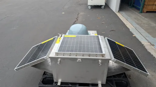 Photo of suitcase-sized steel chest with a solar panel on top and two angled off the sides like wings, and a gray plastic dome mounted on another side.