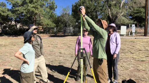 Four students dressed in outdoor sun-protective clothing stand around a tripod supporting an Emlid TRK GNSS receiver. One student attaches the receiver to the tripod. Instructor Beth Pratt-Sitaula provides guidance. The group is on a flat and patchy grass-covered area with trees in the background. 