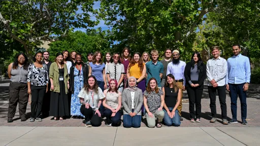 26 NSF NGF interns line up in three rows for a group picture. Five interns kneel in the front on concrete, while the other two rows are interspersed. They are framed by trees in the background.