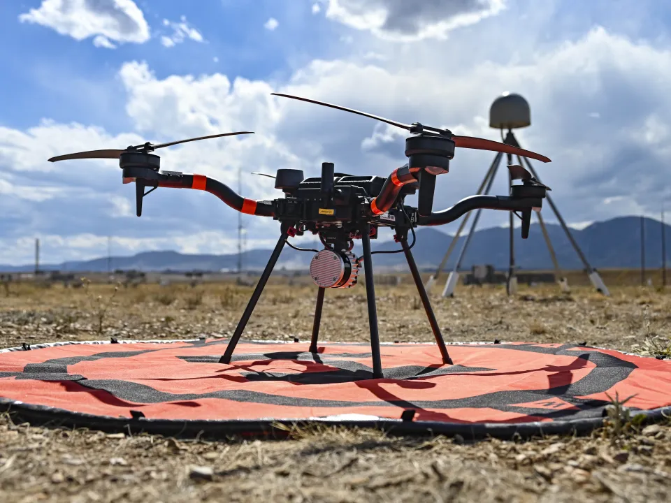 Close photo of a drone on the ground with a GPS station and mountains in the background.