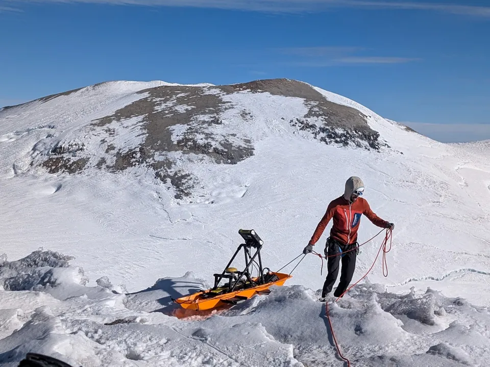 An individual owing a sled with a GPR unit on a snowy mountain landscape.