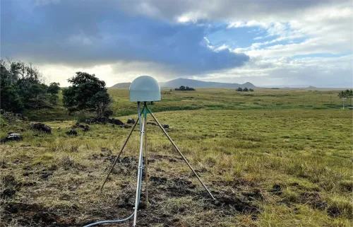 Photo of a permanent GNSS station in a grassy field with trees and clouds in the background.