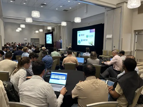 Large conference room with attendees at circular tables looking at projected presentations.