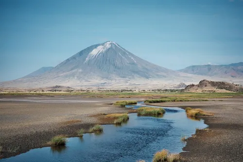 photo of Old Doinyo Lengai, a gray volcano with a patch of streaky white near the summit, foreground is patchy vegetation and a stream