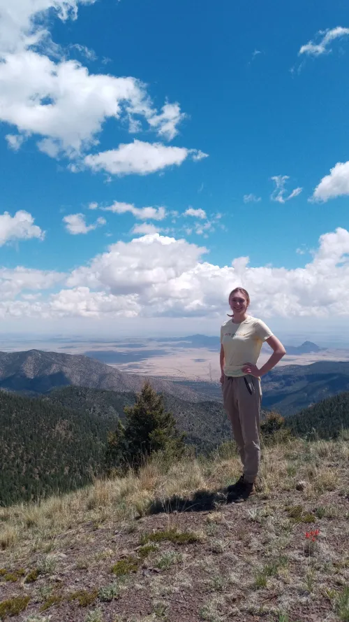 Sarah Ayling posing on Magdalena Ridge with mountains and a blue sky in the background. 