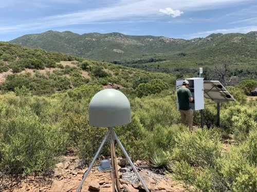 Mario working on a station in the field with green mountains around him.