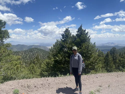 Ann Mariya wearing a baseball hat standing on a hiking path with a mountain range in the background. 