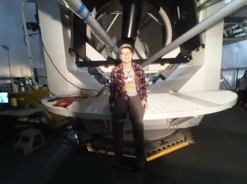 Elliz smiling in front of lab equipment wearing a baseball hat, t-shirt, and red and black flannel. 