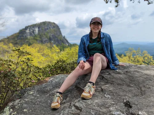 Isabella sitting on a rock in the mountains wearing a blue flannel and a baseball hat.