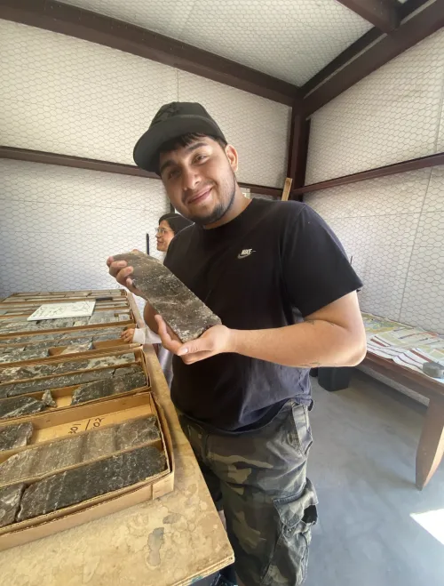 Charles Garcia standing in a lab wearing a black shirt and camo pants holding a slab of rock. 