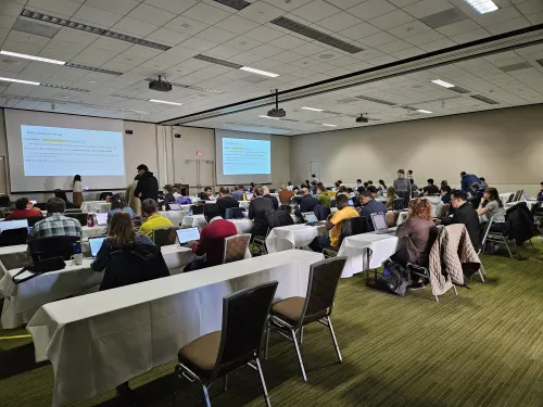 a room full of people at long tables opening up their laptops and looking at projector screens at the front of the room