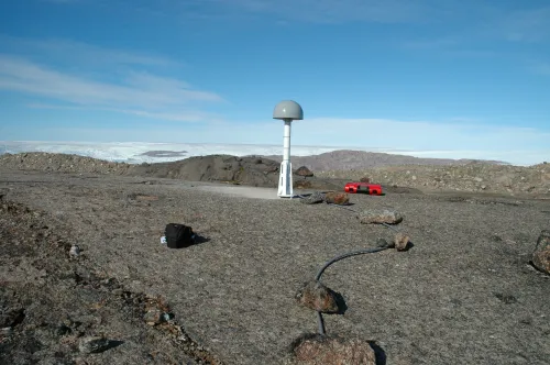 a GPS antenna on a short white metal post affixed to bare bedrock, with white ice marking the horizon in the background