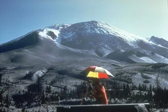 Image of the bulge of Mt. St. Helens in the background, with an onlooker in a bright orange jacket and colorful umbrella in the foreground.