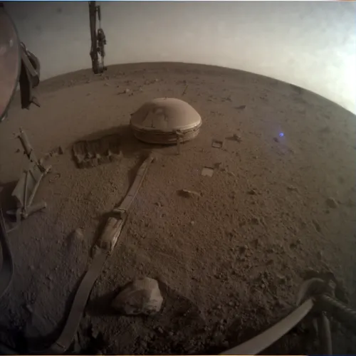 photo of the InSight seismometer sitting on the ground a few feet from the lander body, dusty ground surrounds and the horizon is curved in the fisheye lens view