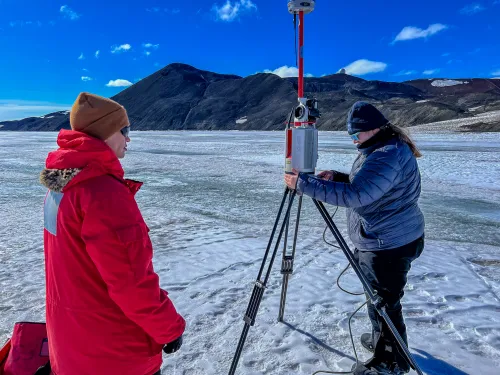 Two people manipulate an instrument on a tripod, standing on sea ice with hills in the distance.