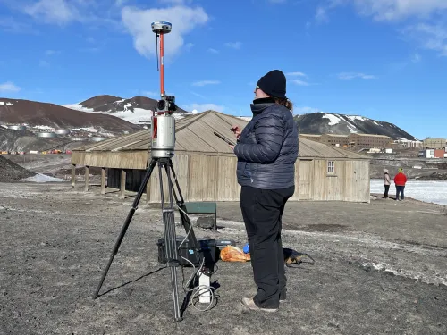 A person stands next to a laser scanner on a tripod, with a square brown hut behind.