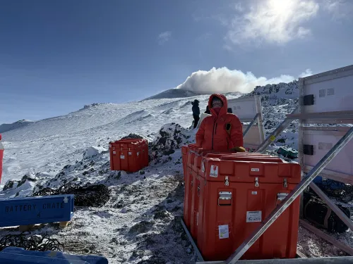 A person stands beside three red cases and two sets of solar panels, with a snowy slope behind and visible steam rising from the peak.