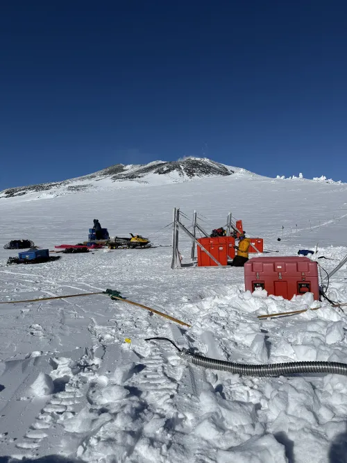 People work around red cases, sets of solar panels, and snowmobiles, with the peak of Erebus in the background.