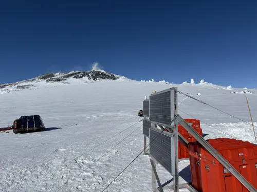 Three red cases and two sets of solar panels on snowy ground, with a helicopter equipment sling on the ground and steaming peak in the background.