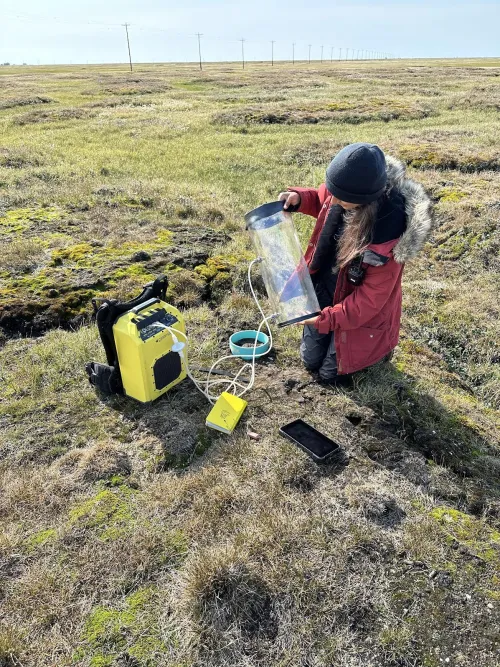 A person kneeling on grassy ground holds a cylindrical chamber connected by tubes to a pump.