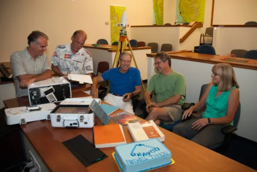 Six people sit and stand around a table during a farewell event for UNAVCO&rsquo;s first GPS receiver, the TI-4100, on July 25, 2014. Vintage GPS equipment and technical manuals are spread across the table, along with a decorated cake that reads &ldquo;UNAVCO Farewell TI-4100.&rdquo; A GPS antenna setup is visible in the background, with topographic maps on the walls.
