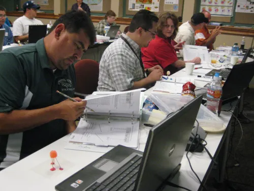 Participants in a UNAVCO Education & Outreach (E&O) short course sit at a long table working with laptops and printed materials. In the foreground, a participant examines a binder while a small gumdrop-and-toothpick model of a GPS station&mdash;nicknamed &ldquo;gumdrop GPS&rdquo;&mdash;sits nearby. Other attendees engage with data sheets and models, illustrating hands-on learning about geodetic motion.