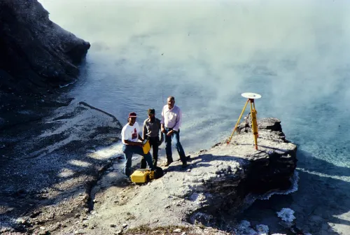 Three geoscientists conduct a GPS survey beside a steaming hydrothermal pool at Yellowstone National Park in 1989. A tripod-mounted GPS antenna is set up on a rocky outcrop. From left to right: Chuck Meertens, Rick Hutchinson, and Bob Smith stand near yellow equipment cases at the water&rsquo;s edge.