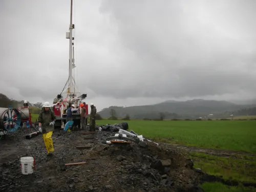 Workers surround a drill rig in a muddy patch surrounded by green grass.