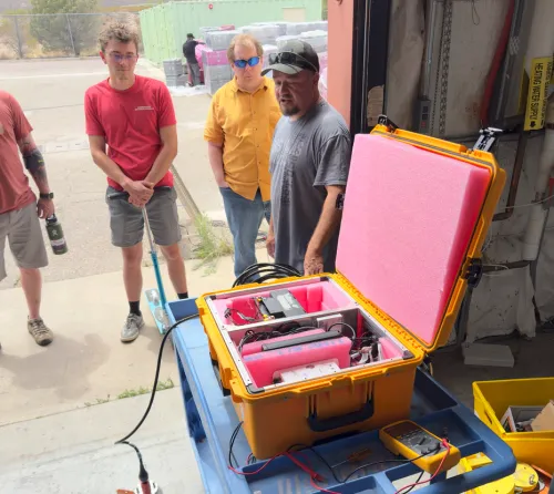 Four engineers surround opened yellow prototype system box that sits on a blue hand cart.