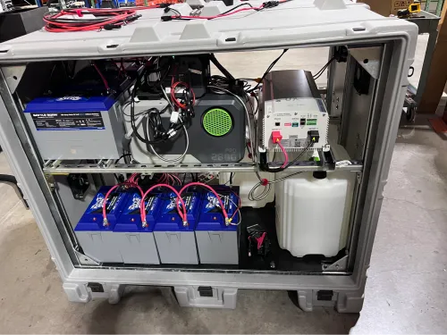 Open view of a similar rugged enclosure displaying the DAS power system components from another angle. Multiple blue battery units are lined up along the bottom, with power electronics, cables, and a compact generator or inverter unit arranged on the shelf above.