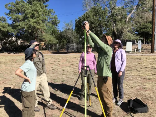 Four students dressed in outdoor sun-protective clothing stand around a tripod supporting an Emlid TRK GNSS receiver. One student attaches the receiver to the tripod. Instructor Beth Pratt-Sitaula provides guidance. The group is on a flat and patchy grass-covered area with trees in the background. 