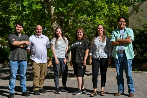 Six people in a row smile at the camera, framed by trees in the background.  