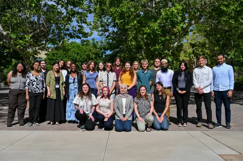 26 NSF NGF interns line up in three rows for a group picture. Five interns kneel in the front on concrete, while the other two rows are interspersed. They are framed by trees in the background.